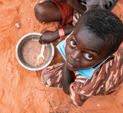 Child on a sandy ground in Luanda with mask and empty bowl, highlighting pandemic challenges.